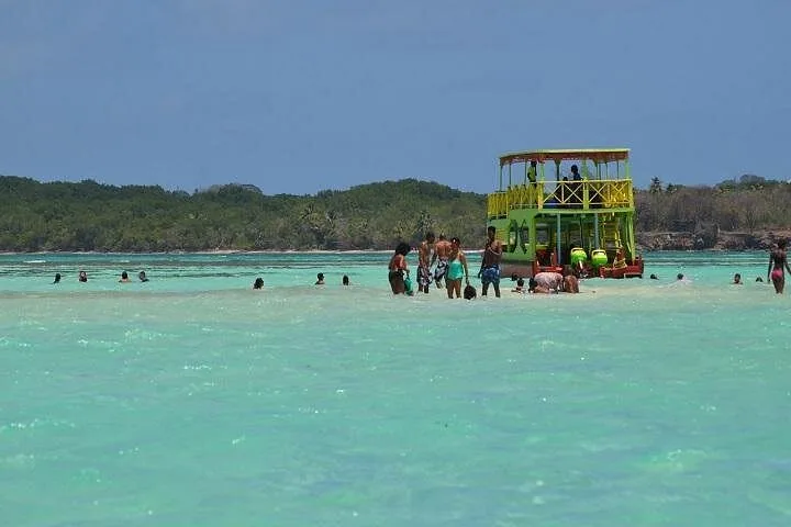 People wading and relaxing around a brightly colored boat in shallow turquoise water.